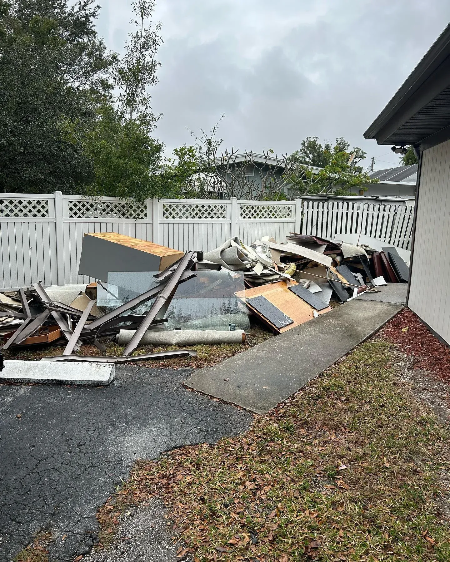 Before view of an outdoor yard with a large pile of wood timber and mixed debris next to a white fence prior to clearance